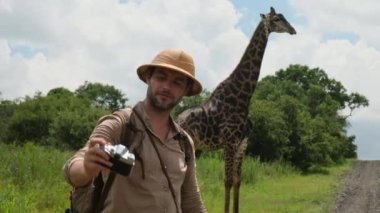Safaride fotoğraf çeken bir adam Afrika savanasında, kurak mevsimde yaprak yiyen vahşi bir zürafa. Safari şapkalı bir erkek gezgin zürafanın selfie çekerken fotoğrafını çekiyor.