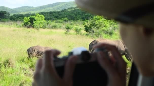 サファリの女性写真家は 乾燥した季節に 赤い地球とアフリカのサバンナの茂みの上で葉を食べる野生のサイを撮影します サファリハットでサイの写真を撮る女性旅行者 — ストック動画