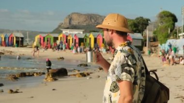 a male traveler in a sun hat drinks hot tea on the ocean against the background of colored houses on the beach. hiker in a shirt with a backpack holds a tourist metal mug with tea and rests