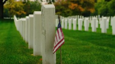 Second World War Cemetery. Military Cemetery Decorated for Memorial Day. American flag with stripes and stars flutters in strong wind. Cemetery graveyard white tombstones at sunset sky 