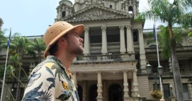 Smiling Hipster man in Glasses with Bag Walking, Looking around at Old City Street. Student, Tourist Concept. Traveling. a man in a safari hat stands near an architectural building in the city center