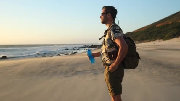 旅行者は岩や波の間の海岸の風景の現代的なカメラで写真を撮る サンセット時に海のビーチで水を飲むバックパック付きの男性旅行者 スポーツや旅行の休暇 — ストック動画