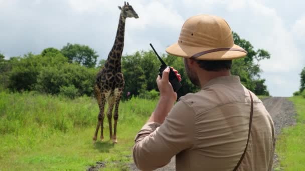 狩猎公园的护林员对着对讲机说话 叫救援队到受伤的动物长颈鹿那里去 非洲国家公园里带着对讲机的偷猎者看着长颈鹿 — 图库视频影像