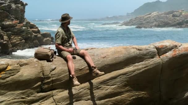 石の上に座ってお茶を飲む男 さまよう冒険の観光 お茶を飲みながら男 日光の下晴れた日に山の上に携帯電話のカメラを持つハイキングバックパッカーの旅行者のキャンパー — ストック動画