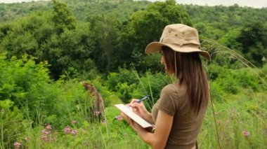 A lone giraffe in the wild of the African savannah walks past a safari car and tourists. a Woman zoologist in a safari hat and a notepad makes notes about wild animals. 