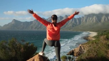 Back view of hipster millennial young woman on top of mountain summit at sunset raises arms into air happy and drunk on life youth and happiness. Watching the sunset with beautiful landscape