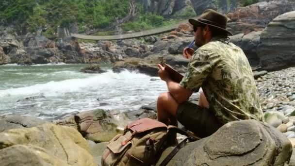 一个戴着帽子和靴子的男性旅行者坐在靠近海洋的岩石上看书 — 图库视频影像