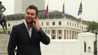 A successful man in a business suit talking on his cell phone outside the South African embassy. A stylish businessman standing in Cape Town with South African flags and a phone in his hands