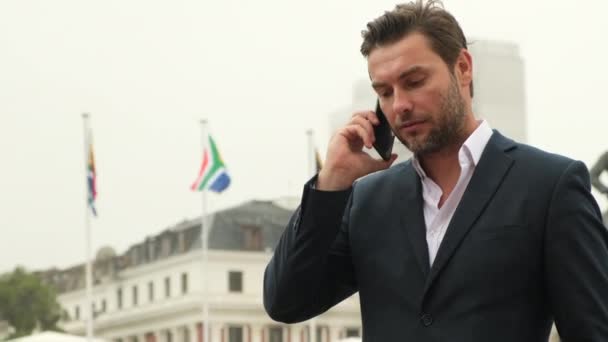 一名穿着西装的成功人士在南非大使馆外面用手机交谈 一位时髦的商人站在开普敦 手里拿着南非国旗和电话 — 图库视频影像