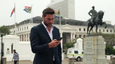 Takım elbiseli başarılı bir adam Cape Town 'daki Güney Afrika bayrağı yakınlarında telefonuyla mesaj atıyor. Şehirde akıllı cep telefonu kullanan yakışıklı bir adam. Akıllı telefondan genç iş adamı.