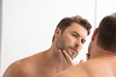 Sakallı seksi adam yüzündeki kırışıklıklara bakıyor. Aynaya bakan adam, hassas cilt, kozmetik tedavisi. Cilt bakımı. neşeli, erkeksi, çekici, çıplak, traşsız, yakışıklı