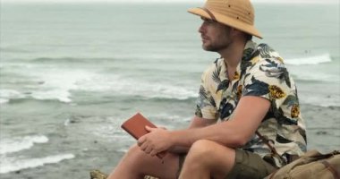  Sırt çantalı, aktif yürüyüş kıyafetleri giymiş turistik bir adam dağ nehri şelalesinin yanında kalıyor ve doğanın tadını çıkarıyor. Not defteri olan bir erkek gezgin dağın zirvesinde oturur ve notlar alır.