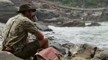 İnsan göl kenarındaki termostan su içer. Sırt çantalı adam aktif yürüyüş kıyafetleri içinde turistik dağ nehri şelalesinin yanında kalıyor ve doğanın tadını çıkarıyor..