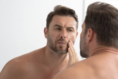 Sakallı seksi adam yüzündeki kırışıklıklara bakıyor. Aynaya bakan adam, hassas cilt, kozmetik tedavisi. Cilt bakımı. neşeli, erkeksi, çekici, çıplak, traşsız, yakışıklı