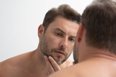 Sakallı seksi adam yüzündeki kırışıklıklara bakıyor. Aynaya bakan adam, hassas cilt, kozmetik tedavisi. Cilt bakımı. neşeli, erkeksi, çekici, çıplak, traşsız, yakışıklı