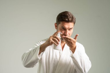 Cilt bakımı kozmetik işlemleri konsepti. Genç ve yakışıklı bir adam mutlu, kendinden emin bir ifadeyle elleri bornozlu ve elinde şampuan şişesiyle yıkanmaya hazırlanıyor.