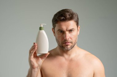 Cilt bakımı kozmetik işlemleri konsepti. Genç ve yakışıklı bir adam mutlu, kendinden emin bir ifadeyle elleri bornozlu ve elinde şampuan şişesiyle yıkanmaya hazırlanıyor.