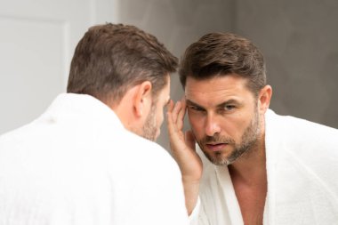 Banyoda beyaz bornozlu bir adam yüz spa 'sındaki deriye bakıyor. Cilt bakımı için cilt bakımı ürünü. Cilt bakımı. Erkek yüz kremi. Mükemmel yumuşak ciltli bir adam. Güzellik ürünleri.