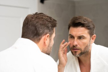Banyoda beyaz bornozlu bir adam yüz spa 'sındaki deriye bakıyor. Cilt bakımı için cilt bakımı ürünü. Cilt bakımı. Erkek yüz kremi. Mükemmel yumuşak ciltli bir adam. Güzellik ürünleri.