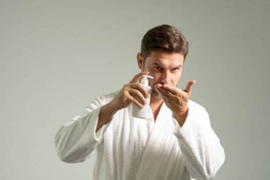 Cilt bakımı kozmetik işlemleri konsepti. Genç ve yakışıklı bir adam mutlu, kendinden emin bir ifadeyle elleri bornozlu ve elinde şampuan şişesiyle yıkanmaya hazırlanıyor.