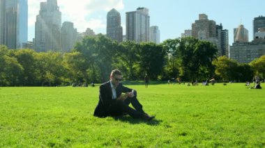  Parkta rahatlayan yakışıklı iş adamının portresi. Şehir parkında iş adamı öğle yemeği molası veriyor. New York 'taki Central Park' ta takım elbiseli Amerikalı bir işadamı çimlerin üzerine oturup mesaj yazıyor.