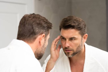 Banyoda beyaz bornozlu bir adam yüz spa 'sındaki deriye bakıyor. Cilt bakımı için cilt bakımı ürünü. Cilt bakımı. Erkek yüz kremi. Mükemmel yumuşak ciltli bir adam. Güzellik ürünleri.