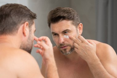 Adam banyoda nemlendirici yüz kremi kullanıyor. Krem tutan mutlu genç adam. Yüzüne krem süren 40 'lı yaşların portresi. Yakışıklı ciddi adam taze cilt kremi sürerek eğleniyor..