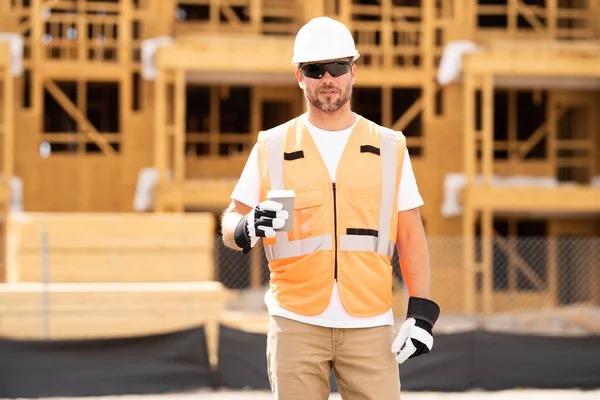 Coffee, engineer and construction worker relax on break at construction site. handsome male builder in hard hat smiling at camera. Construction Worker on Duty. Contractor and the Wooden House Frame