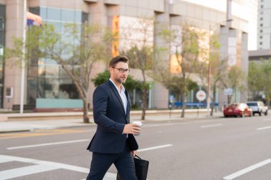 Sırt çantalı bir iş adamı şehirde yürüyor. Şık takım elbiseli erkek girişimcinin yan görünüşü ve sabah işe giderken yaya geçidinde yürürken içkiyle birlikte yürümesi. Sokaktaki iş adamı