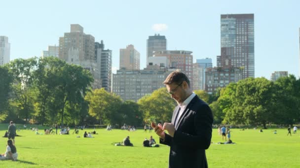 公園でリラックスしたハンサムなビジネスマンの肖像画 ニューヨークのセントラルパークのスーツでアメリカのビジネスマンを昼食をとるビジネスマンが草の上に座ってテキストメッセージを入力 — ストック動画