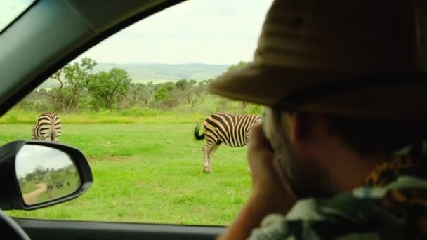一名头戴狩猎帽的男性游客在非洲大草原拍摄动物的照片 一名男性旅行者坐在一辆狩猎车上 为一只国家保护区斑马拍摄野生动物的照片 — 图库视频影像