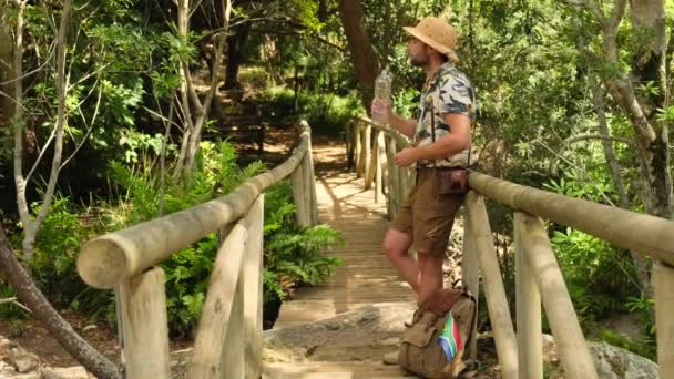 ショートパンツとシャツの男性観光客は ジャングルのプラスチックボトルから水を飲む ハイカーマンは山を長いトレッキングの後 プラスチックボトルから水で渇きを癒します — ストック動画