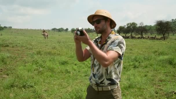 アフリカのキリンと一緒にサファリを旅行するカメラを持つ男性の観光客 サバンナで大量のキリンが 草を食べている サファリの服の男が野生動物の写真を撮る — ストック動画