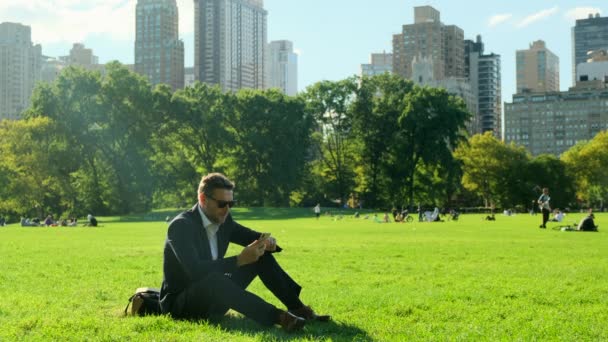 一个英俊的商人在公园里放松的画像 在纽约中央公园 一位身穿西装的美国商人坐在草地上发短信 他正在午休 — 图库视频影像