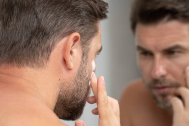 Sakallı bir adam yüzüne krem sürüyor, alnına, saçına, burnuna, gözlerine, ağzına ve boynuna odaklanıyor..