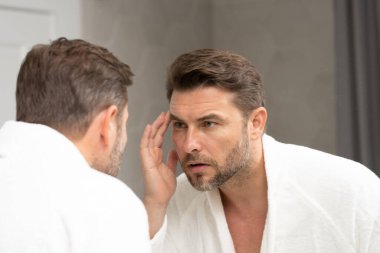 Banyoda beyaz bornozlu bir adam yüz spa 'sındaki deriye bakıyor. Cilt bakımı için cilt bakımı ürünü. Cilt bakımı. Erkek yüz kremi. Mükemmel yumuşak ciltli bir adam. Güzellik ürünleri.