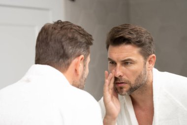 Banyoda beyaz bornozlu bir adam yüz spa 'sındaki deriye bakıyor. Cilt bakımı için cilt bakımı ürünü. Cilt bakımı. Erkek yüz kremi. Mükemmel yumuşak ciltli bir adam. Güzellik ürünleri.
