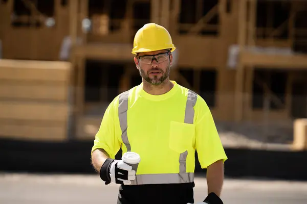 A construction worker at a construction site drinks coffee during a break. builder man in a helmet and vest holding a glass of coffee. a construction inspector in a helmet drinking coffee.