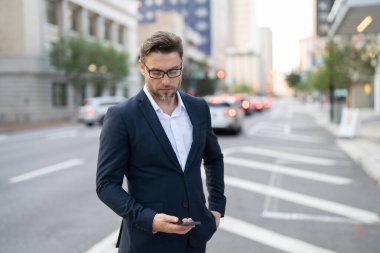 Yakışıklı genç bir işadamı telefonunu kullanıp sokakta dikiliyor. Yakışıklı, gülümseyen genç iş adamı şehrin dışında cep telefonu kullanarak şık bir şekilde dikiliyor.