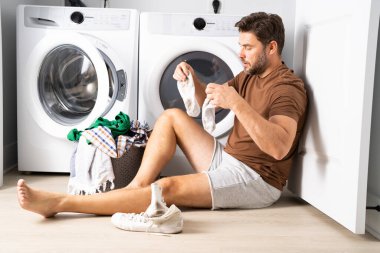 Çamaşır odasında çamaşır makinesinin yanında giysileri sıralayan mutlu bir adam. Adam kirli çamaşırlarıyla çamaşır makinesinin yanında bir evin zemininde oturuyor. Bir adam çamaşırlarını evde yıkıyor. Kirli çamaşırları olan bir adam.