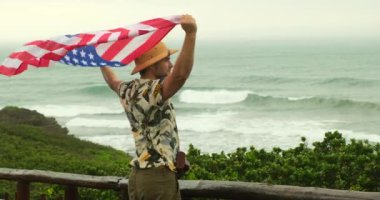 Okyanus kıyısındaki bir dağın kenarında elinde Amerikan bayrağı olan bir erkek gezgin. Amerikan bağımsızlık gününde Amerikan bayrağını gururla taşıyan tişörtlü bir Amerikalı.