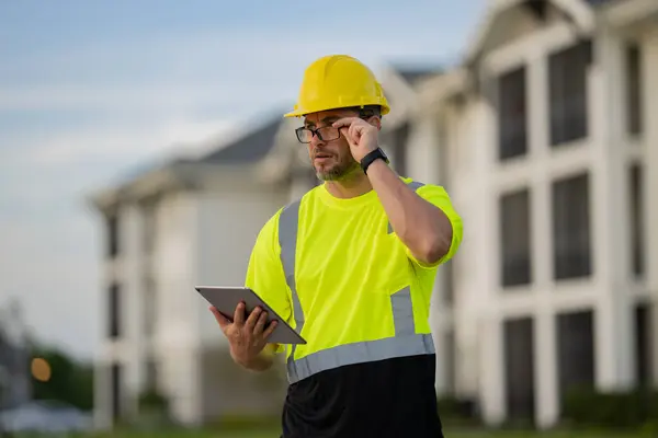 stock image A man in a yellow safety vest is looking at a tablet while wearing a hard hat. He is focused on the device, possibly checking for safety information or instructions