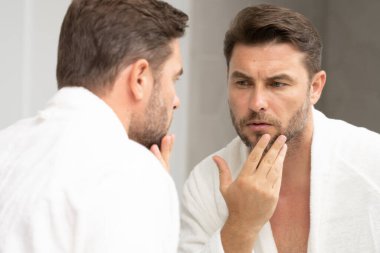 Banyoda bornozlu bir adam yüzündeki kırışıklıklara ve sivilcelere bakıyor. Bir adam aynadaki yansımasına bakıyor. Bir adam aynadaki yansımasına bakıyor.. 