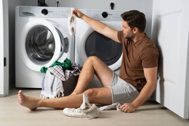 Çamaşır makinesinin yanında çamaşır sıralayan bir adam. Kirli çamaşırları olan bir çamaşır makinesinin yanında bir adam oturuyor. Çamaşır makinesinin yanında kirli bir çorap tutan bir adam, kirli ayakkabılar, kuru temizlemeci..