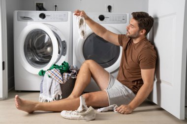 Çamaşır makinesinin yanında çamaşır sıralayan bir adam. Kirli çamaşırları olan bir çamaşır makinesinin yanında bir adam oturuyor. Çamaşır makinesinin yanında kirli bir çorap tutan bir adam, kirli ayakkabılar, kuru temizlemeci..