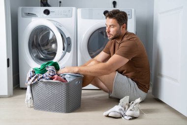 Bir adam banyonun zemininde oturuyor, kirli çamaşırları gri bir sepetten ayırıyor. Elbiselerinden birini çıkarmak için bir çamaşır makinesine uzanıyor. Banyo temiz ve modern. 