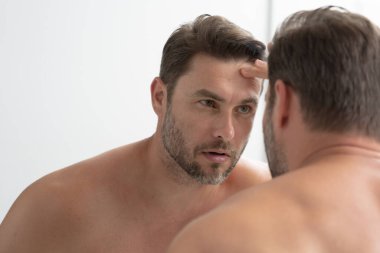 Banyoda bornozlu bir adam yüzündeki kırışıklıklara ve sivilcelere bakıyor. Bir adam aynadaki yansımasına bakıyor. Bir adam aynadaki yansımasına bakıyor.. 