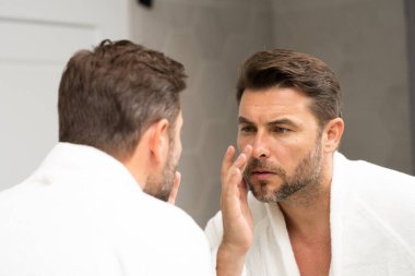 Banyoda bornozlu bir adam yüzündeki kırışıklıklara ve sivilcelere bakıyor. Bir adam aynadaki yansımasına bakıyor. Bir adam aynadaki yansımasına bakıyor.. 