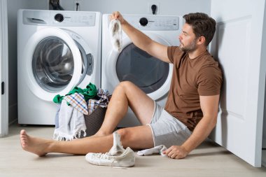 Çamaşır odasında çamaşır makinesinin yanında giysileri sıralayan mutlu bir adam. Adam kirli çamaşırlarıyla çamaşır makinesinin yanında bir evin zemininde oturuyor. Bir adam çamaşırlarını evde yıkıyor. Kirli çamaşırları olan bir adam.