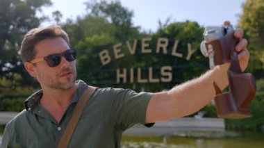 Klasik film kamerasında fotoğraf çeken bir adam. Retro kameralı şık erkek turist, Los Angeles 'taki Beverly Hills tabelasının önünde fotoğraf çekiyor.